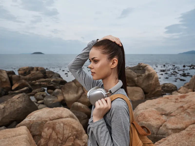 Young woman enjoying music by the sea, wearing casual grey outfit, holding headphones, looking thoughtfully at the horizon, serene coastal background
