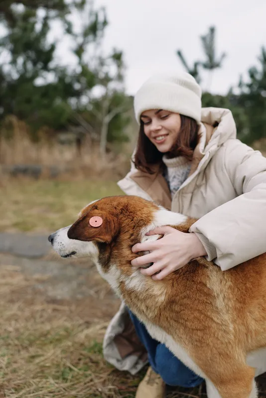 Woman in a warm winter coat and beanie cuddling a dog outdoors in a park, close interaction and gentle touch, conveying warmth, care and companionship.