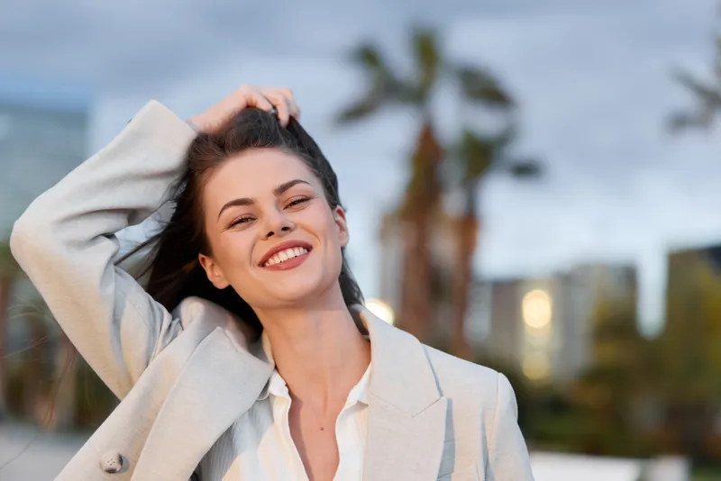 Young woman smiling confidently in beige business jacket outdoors with cityscape and palm trees in soft focus background bright daylight