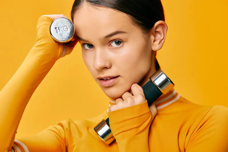 Woman in vibrant orange athletic wear with dumbbells resting on shoulders, ready for workout, focused expression, bright orange studio backdrop and energetic fitness mood.