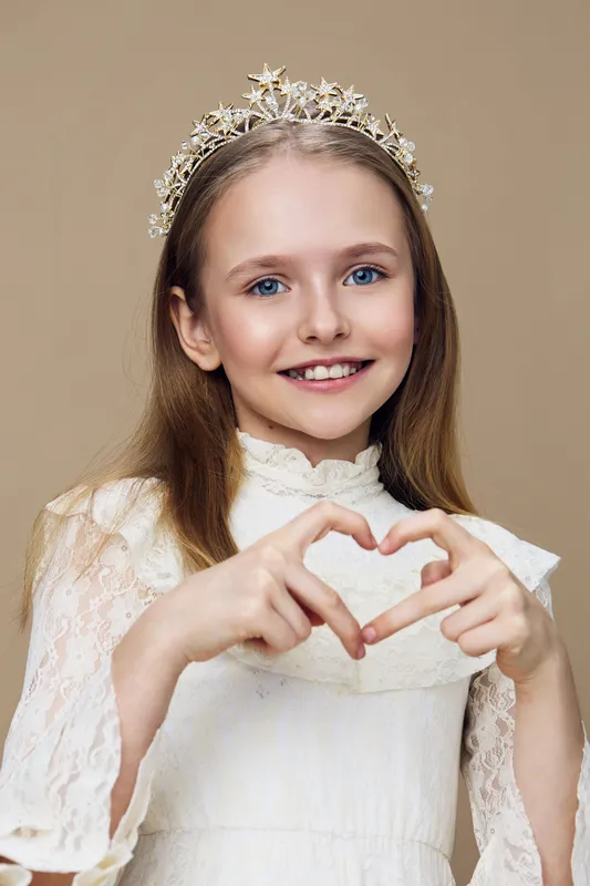 Young girl wearing a crown, making a heart shape with her hands, smiling joyfully against a soft beige background