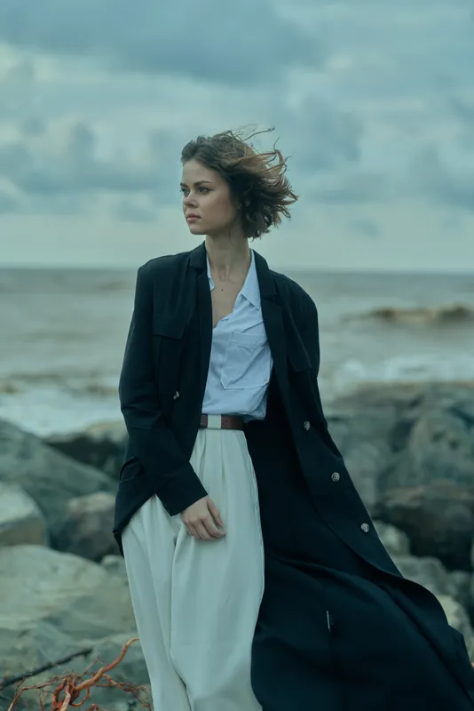 Woman in a long navy coat over a light dress stands on a rocky coastline, wind ruffling her hair, ocean and cloudy sky behind, a moody fashion moment by the sea.