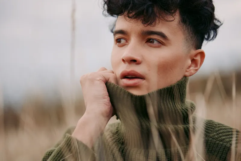 A close portrait of a young person in a thick olive knit turtleneck, conveying authenticity through subtle expression and quiet intensity in a natural outdoor setting, and veracity.
