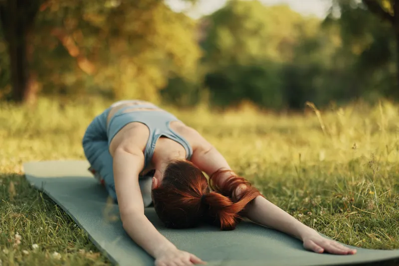 Young woman practicing yoga outdoors, demonstrating a stretching pose on a mat in a serene green environment, reflecting wellness and tranquility