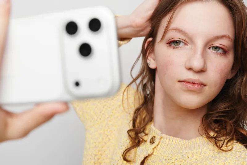Young woman with curly hair wearing yellow sweater takes selfie with smartphone in bright studio. Concept of social media, self portrait, and casual lifestyle photography.