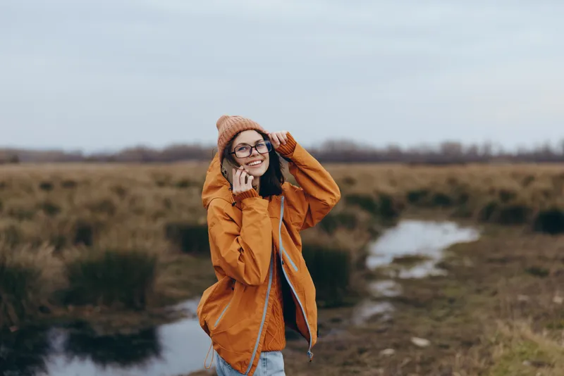 Woman outdoors field river coat hat smile glasses winter lifestyle, smiling and adjusting knit hat, orange coat on woman, rural countryside landscape, natural daylight creates calm mood