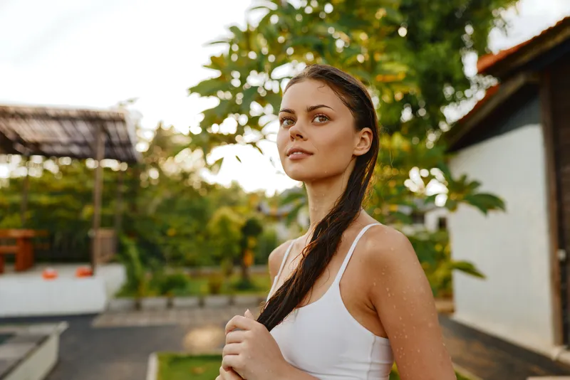Young woman with long braided hair stands outdoors in a white tank top during golden hour, looking confidently into the distance in a relaxed lifestyle scene.