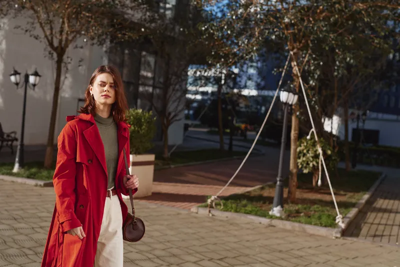 Young woman with red hair wearing a red trench coat and white pants stands outdoors on a sunny autumn day with a thoughtful expression.