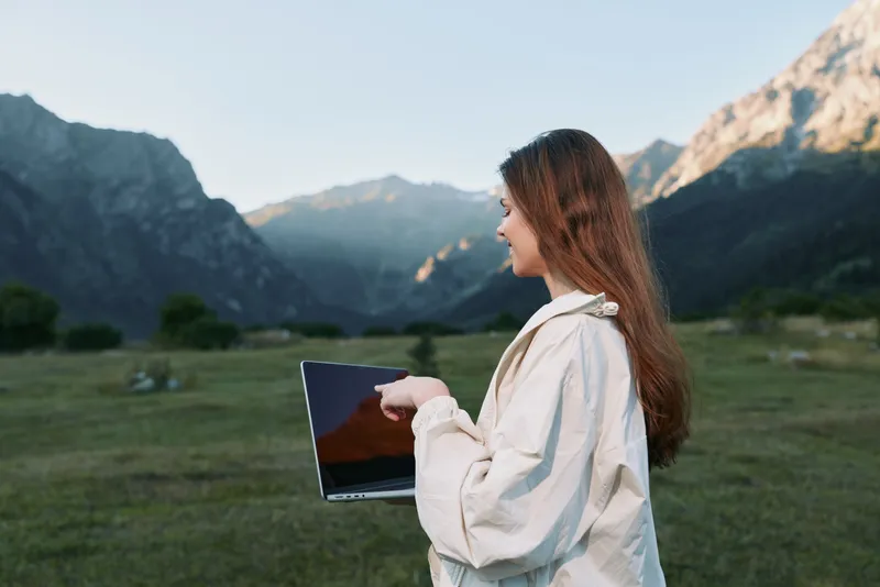 A thoughtful woman stands outdoors in a scenic valley, holding a laptop and gazing toward distant peaks. The scene conveys focus, exploration, and digital work in nature.