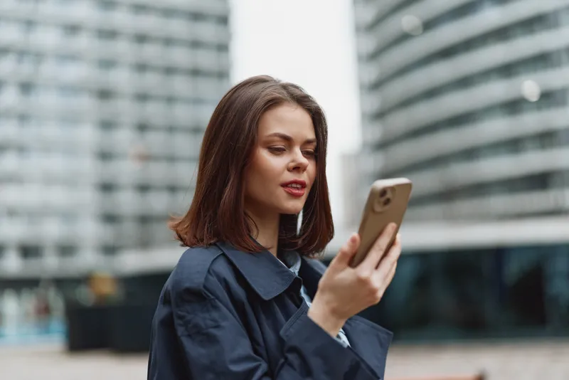 Young woman with straight brown hair wearing a dark coat looks at her smartphone with a slight smile in an urban environment with modern buildings in the background.