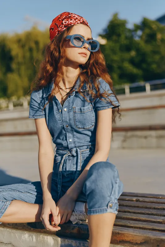 Lifestyle woman in modern boho-western denim street style wearing blue jumpsuit, red bandana, and sunglasses with relaxed, casual outdoor mood and film color tone.