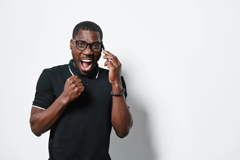 African American man with dark skin wearing glasses and black polo shirt talking on phone and celebrating success with fist pump on white background in studio.