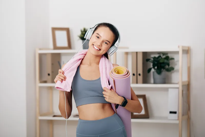 Fitness woman in a gray sports bra and leggings with headphones, towel over shoulders, holding a yoga mat in a bright home gym for a dynamic workout and wellness moment.