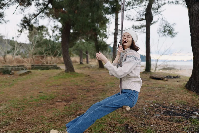 Woman on a rope swing in an outdoor park, laughing and enjoying nature. Cozy sweater, jeans, pine trees by a lakeside, capturing a carefree moment and playful mood suitable for lifestyle stock