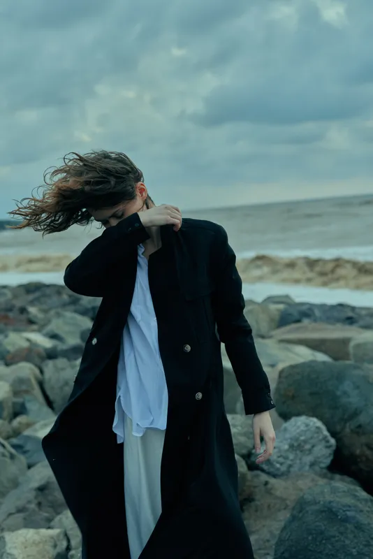 Windy beach scene with flowing hair, a long black coat, white shirt, and rocky shore as sea and dramatic sky create a bold outdoor fashion mood.