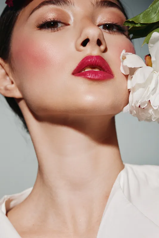 Close-up of a fashion woman with flawless skin and glossy red lips posing elegantly holding white flower near face studio shot beauty creative makeup concept.