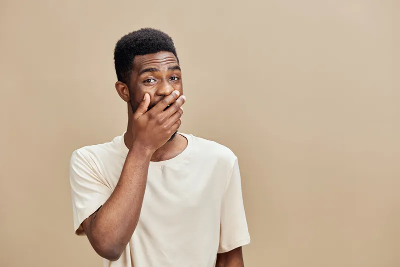 Young African man covering mouth with hand, expressing surprise or embarrassment, wearing a light t shirt against a neutral background