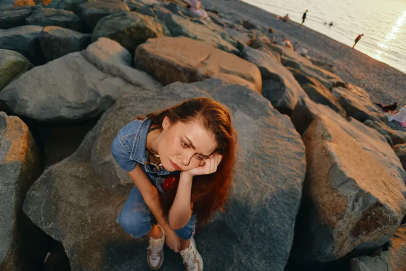 thoughtful woman sitting on large rocks near water during sunset in casual denim outfit with long red hair and relaxed mood