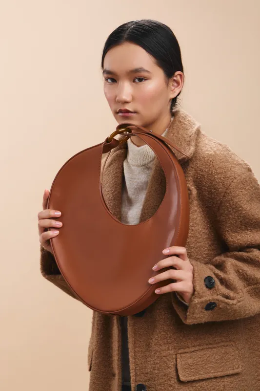 asian woman holds a round brown bag in a neutral studio setting. She wears a tan coat over a light sweater, presenting a calm, stylish fashion moment.