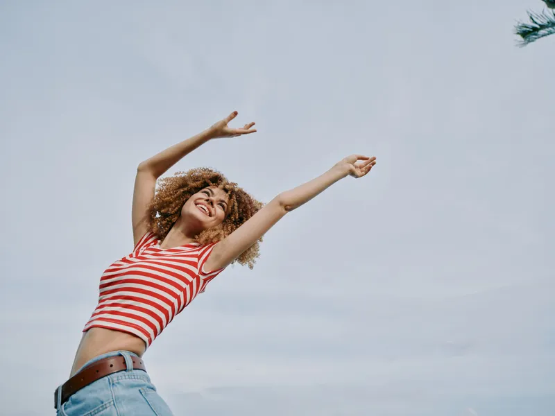 A joyful woman with curly hair raises her arms in a carefree pose, wearing a red and white striped top and denim jeans against a bright blue sky.
