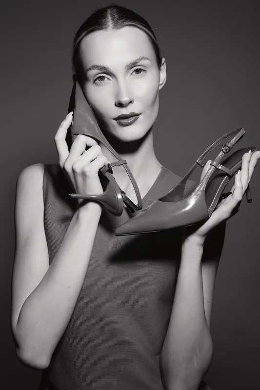 Elegant white woman with fair skin holds high heel shoe near face in black and white studio portrait. Concept for fashion, style, beauty, and footwear accessories.