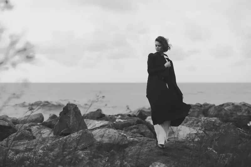 Woman in a flowing black dress stands on rocky shoreline beside the sea, wind catching fabric, moody monochrome scene emphasizing solitude, elegance and nature's rugged beauty