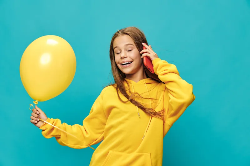 Girl in yellow hoodie holding yellow balloon and talking on smartphone with eyes closed on blue background for joyful communication and celebration.