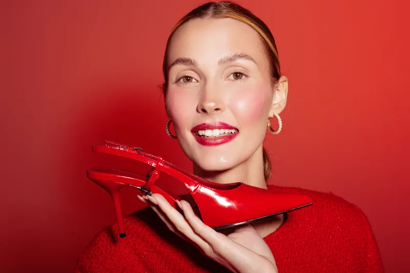 Smiling woman in red sweater holds red high heel shoe near face on red background. Fashion concept with bright colors, style, and beauty in studio lighting for footwear promotion.