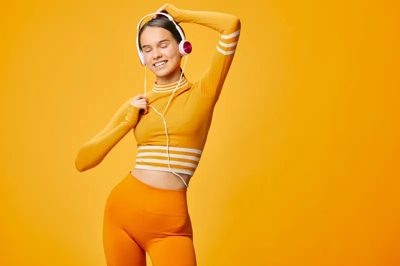 Fitness sport woman in vibrant orange activewear dances with headphones, showing energy and confidence in a bold studio. Dynamic pose captures movement, balance and style for a sporty lifestyle