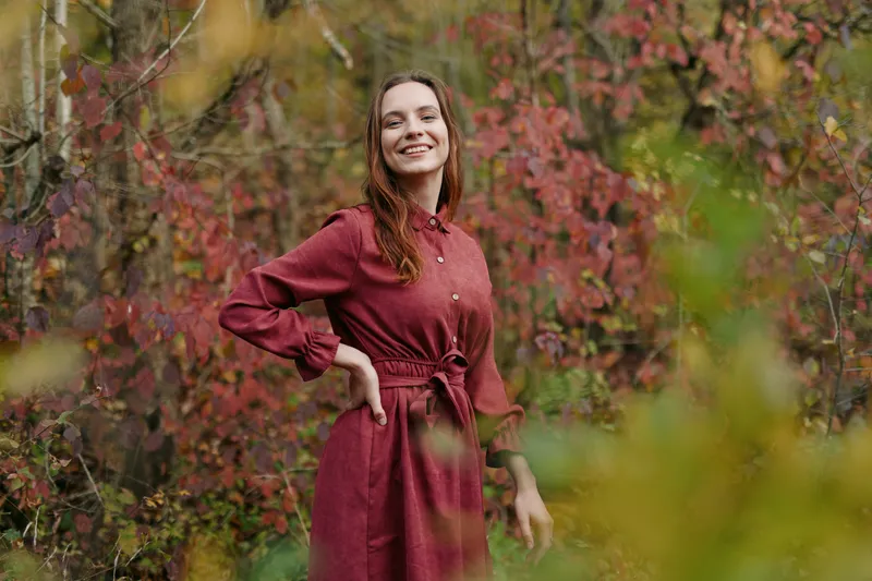 A genuine portrait of a smiling woman in a deep red dress, posed with hands on hips, conveying candid confidence and natural authenticity in a warm outdoor setting.