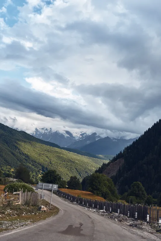 Mountain road winding through green valley with forested slopes under dramatic clouds sky, rural fence and distant peaks, scenic nature landscape view