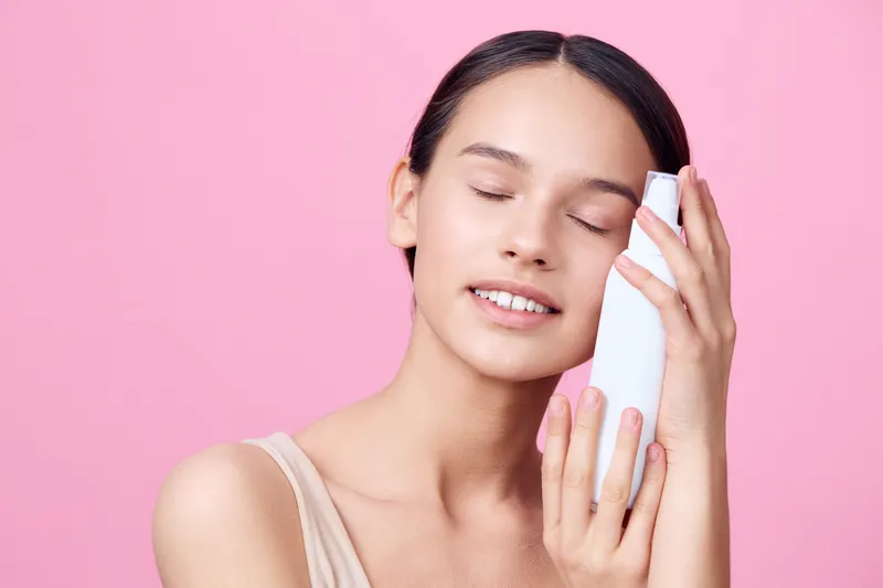 Young woman holding a skincare product, smiling gently on a soft pink background, radiating happiness and self care Ideal for beauty and wellness concepts