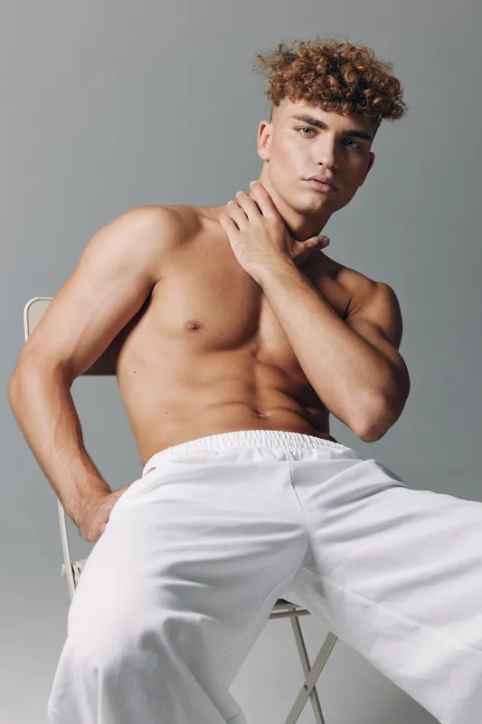 Young muscular man shirtless sitting in white pants on chair with hand on neck for workout bodystudy and fitness strength with intense thoughtful expression in studio.