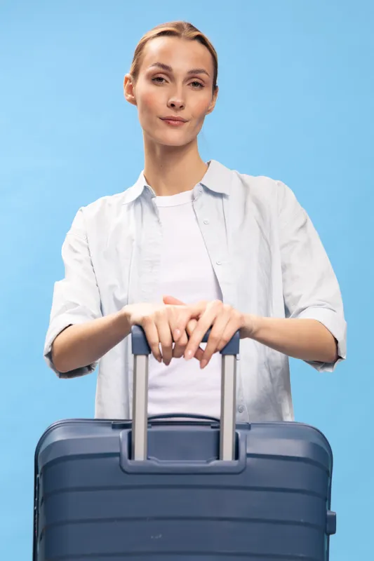 Young woman with suitcase standing against blue background, wearing casual white shirt. Concept of travel, vacation, and tourism with confident and relaxed mood.