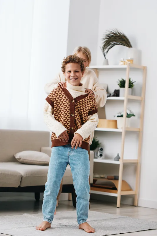 Bright living room scene features a smiling child in knitted cardigan and denim jeans, with a supportive adult gently embracing from behind in a warm family moment.