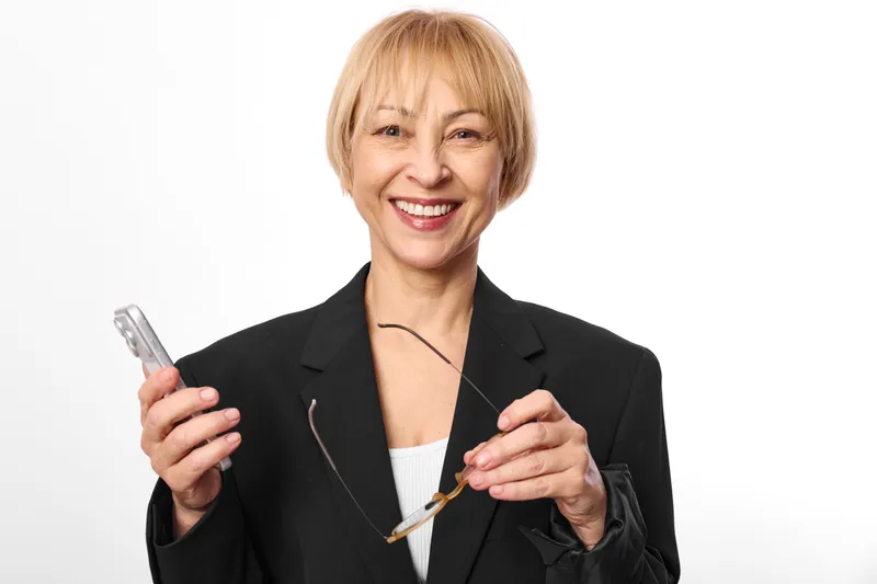 Smiling mature woman in business jacket holding smartphone and pen, corporate portrait on clean white background, confident expression, studio lighting, modern communication