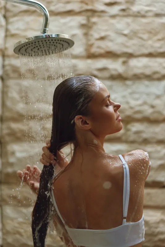 young woman washing long hair under rainfall shower with stone wall background, wellness, relaxation, wet skin, self-care moment