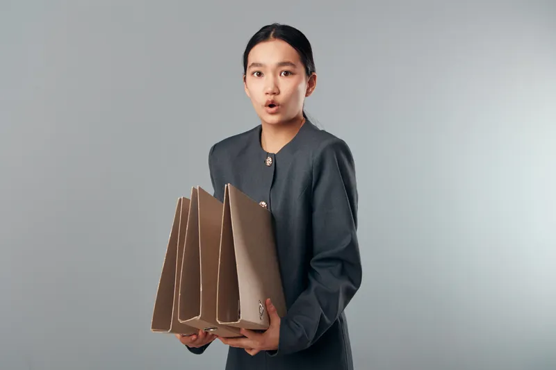 Young woman in gray jacket holding multiple cardboard folders with surprised expression on gray background. Female office worker in casual outfit with documents for organization or presentation.