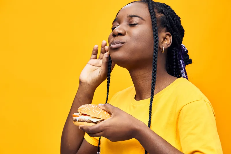 Young Black woman enjoying a delicious burger against a vibrant yellow background, expressing joy and satisfaction, representing a celebration of good food and flavors