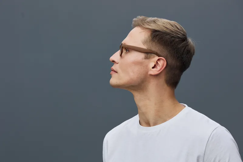 Close-up profile of a young man with glasses and neatly styled brown hair wearing casual white t-shirt, looking sideways, isolated on plain dark background, studio portrait, modern lifestyle concept.