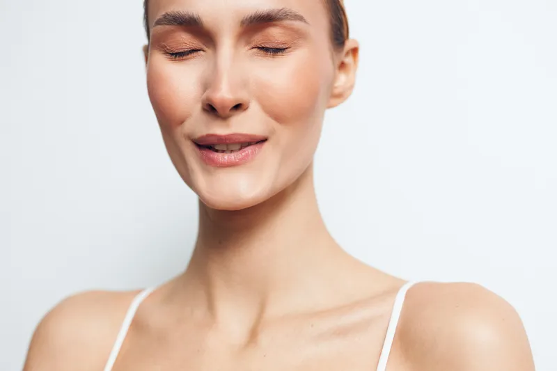 Young woman with closed eyes and relaxed face enjoys calm moment in minimal white top isolated on light background, expressing serenity and natural beauty with smooth skin.