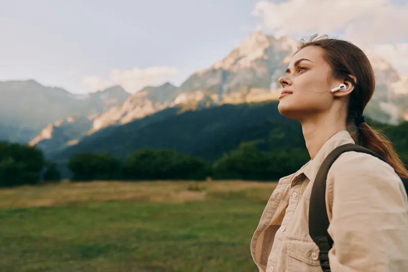 Young woman with long hair enjoys a peaceful moment outdoors with wireless earbuds in a mountainous landscape during a clear day.