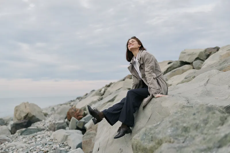 Young woman in casual outfit sits on large rocks by the shore, enjoying peaceful moment under cloudy sky. Concept of relaxation, nature, and outdoor lifestyle near the sea.