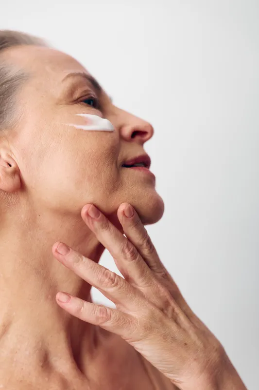 Mature woman applies white facial cream on cheek with fingers. Close-up side view with soft lighting on neutral background. Concept of skincare, aging skin care, and self-care routine.