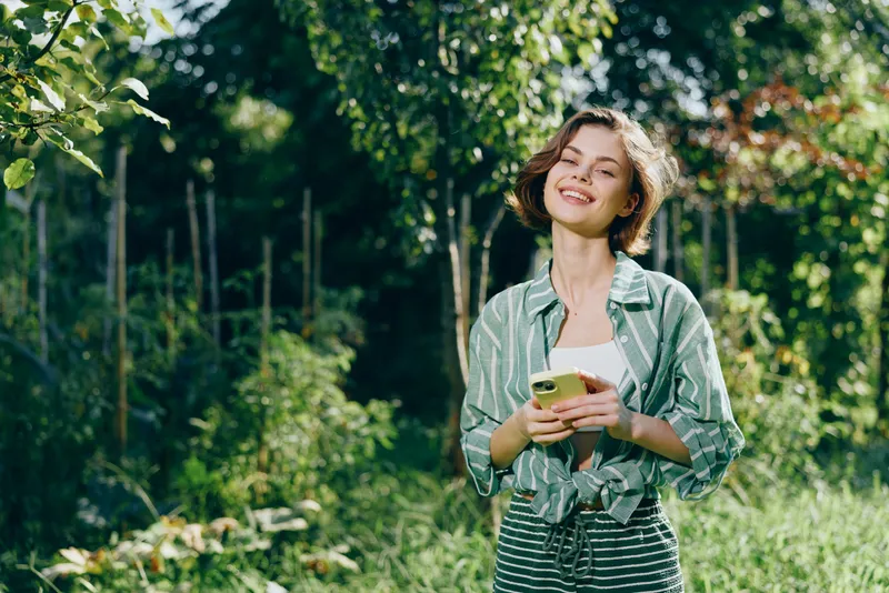 Cheerful young woman standing in a sunny garden, holding a smartphone with a bright smile, wearing a green striped shirt and casual dress.