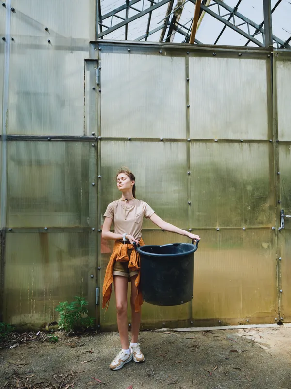 woman greenhouse bucket gardening plants young casual seedlings potting in urban conservatory, female gardener in shorts and sneakers holding large black container for soil and planting