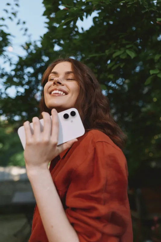 A young woman stands outdoors in a park, smiling while holding a smartphone. A casual outfit in red tones and natural light convey a carefree lifestyle and candid outdoor portrait
