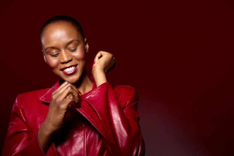 Confident black woman with dark skin wearing a red leather jacket smiles with closed eyes on a dark red background. Concept of fashion, beauty, elegance, and self-confidence.