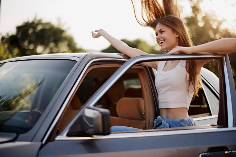 Young woman enjoying a road trip with her hair blowing in the wind from a car window, capturing the essence of freedom and adventure in a sunny outdoor setting.