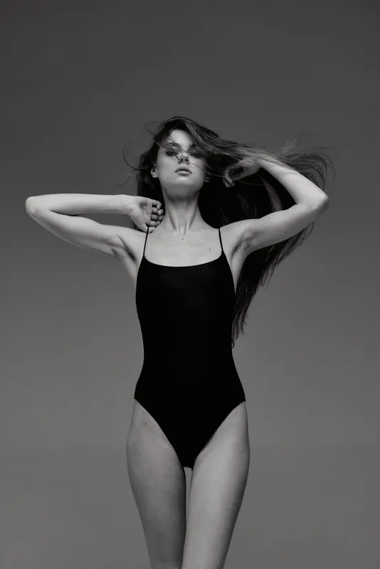 Black and white portrait of a slender woman in swimsuit posing with hands in hair on a plain background.